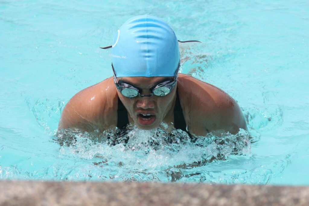 A pool swimmer performing competition breaststroke wearing a blue hat. While accessible, breaststroke for triathlon in an inefficient way to swim.