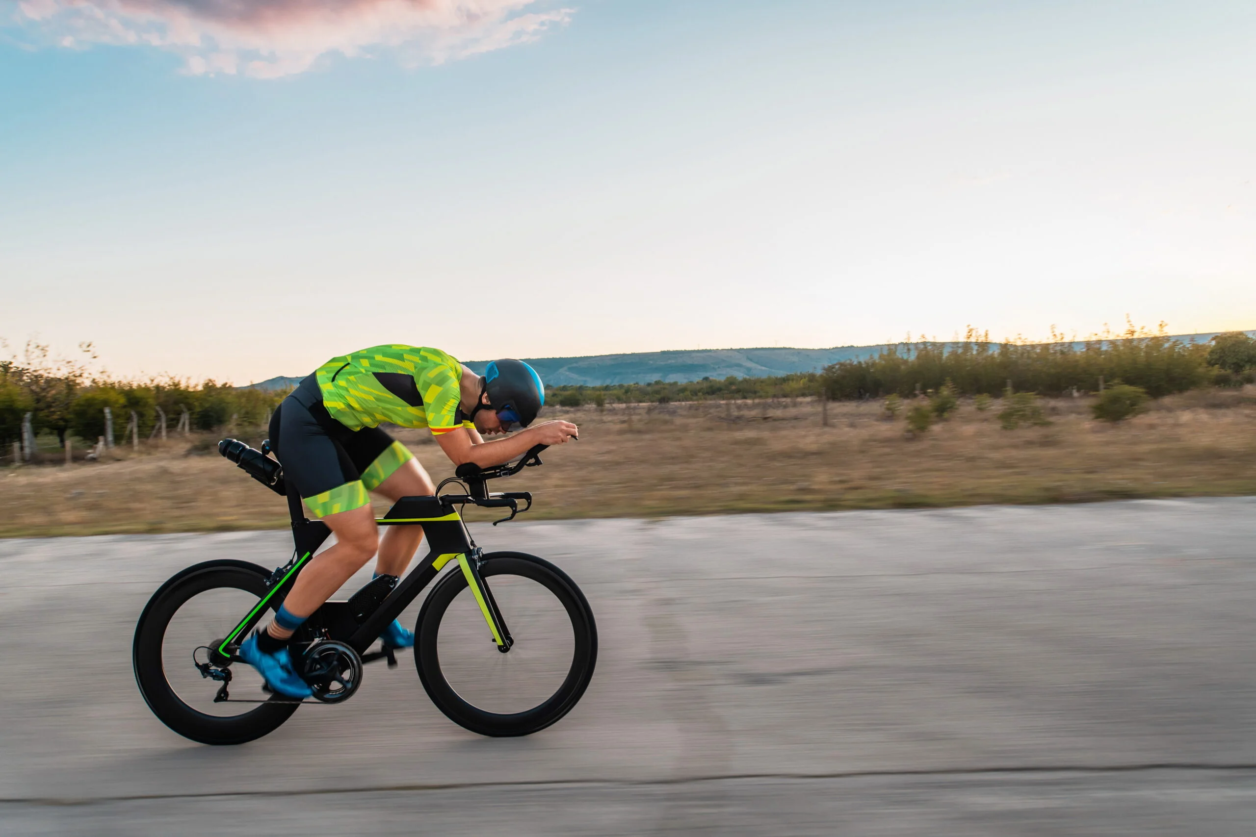 Triathlete riding his bicycle during sunset, preparing for a marathon. The warm colors of the sky provide a beautiful backdrop for his determined and focused effort